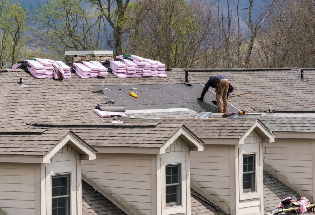 A worker repairs a shingled roof with stacks of roofing materials nearby.