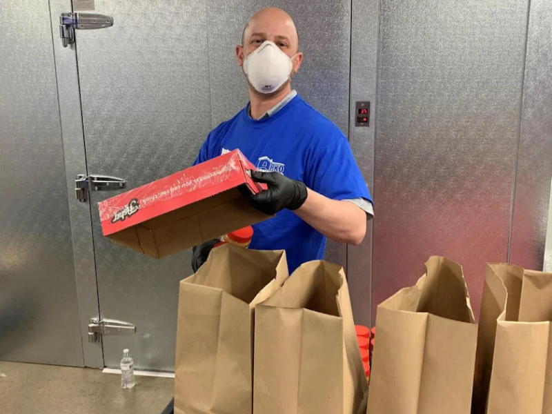 Man wearing a mask and gloves packing items into brown paper bags inside a metal-walled room.