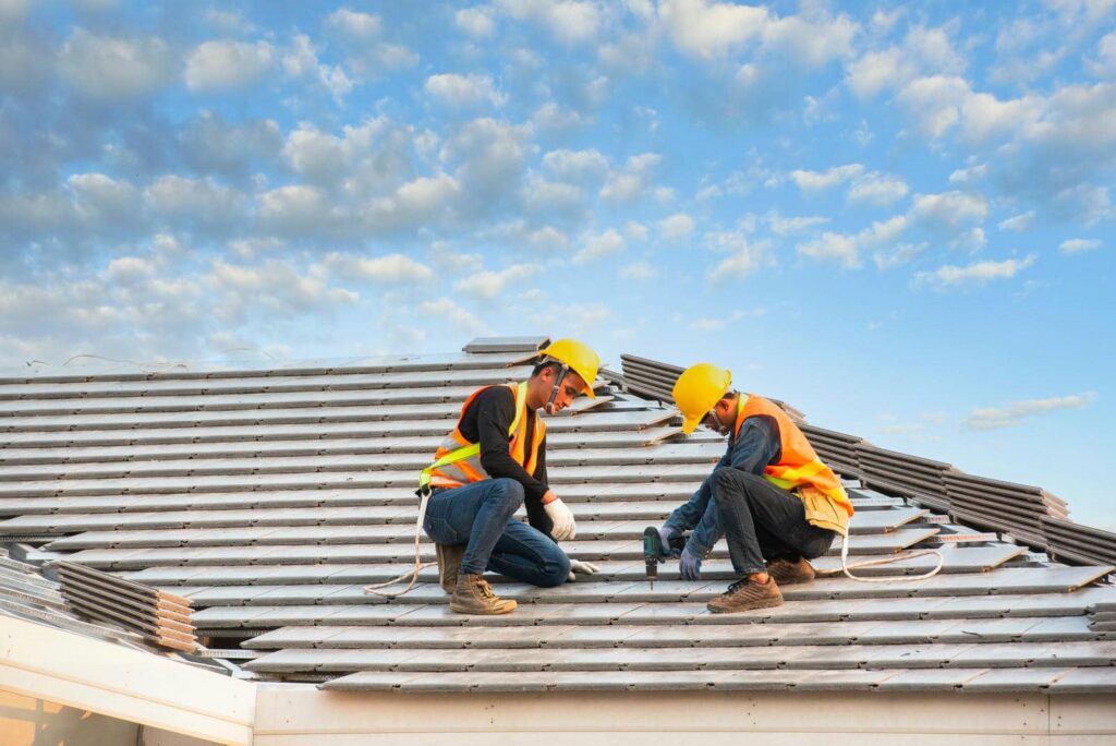 Two construction workers in safety gear installing roof tiles under a partly cloudy sky.