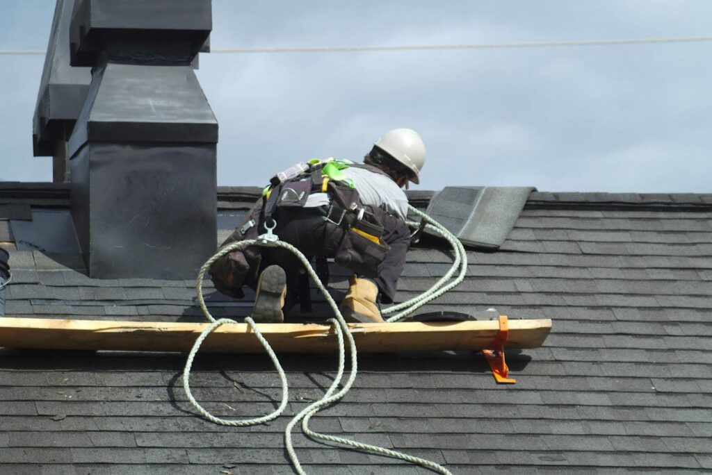 Roofer wearing a safety harness works on a shingled roof near a chimney.