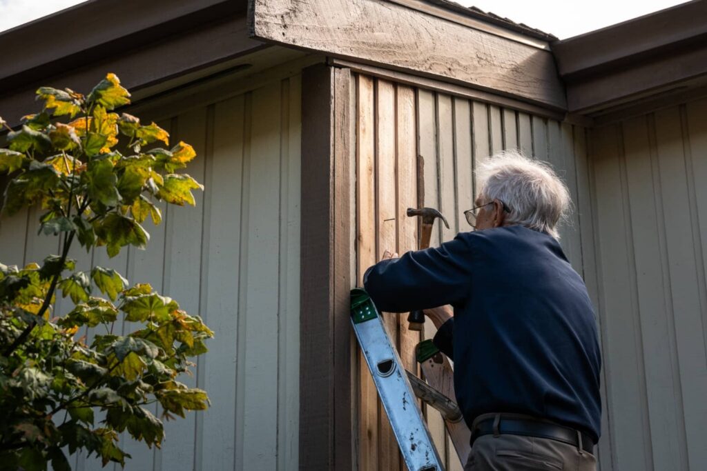 Older man on a ladder hammering nails into wooden siding of a house.