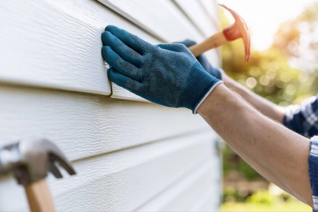 Hands wearing blue gloves installing white vinyl siding on a house with a hammer.
