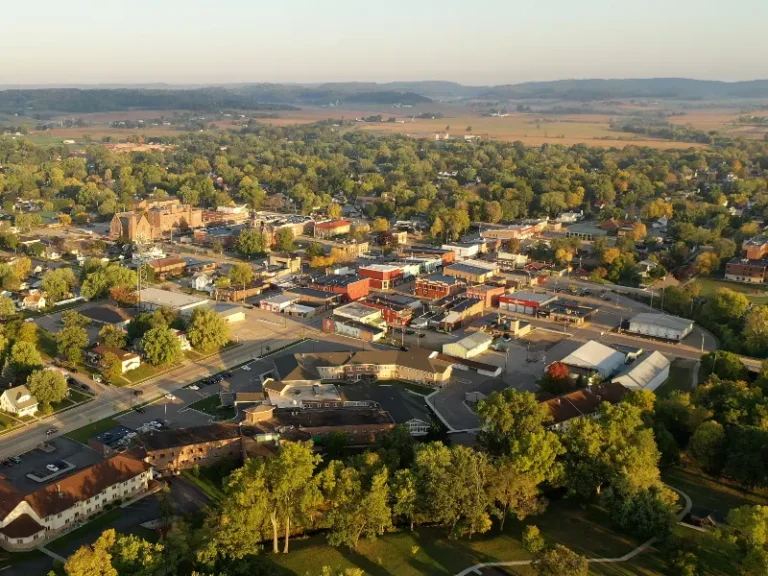 Aerial view of a small town with buildings, trees, and distant hills under a clear sky.