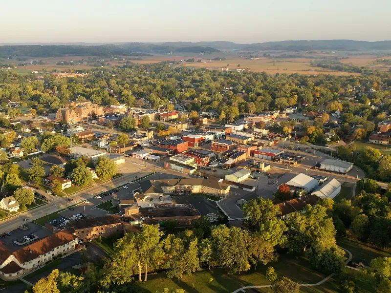 Aerial view of a small town with buildings, trees, and distant hills under a clear sky.