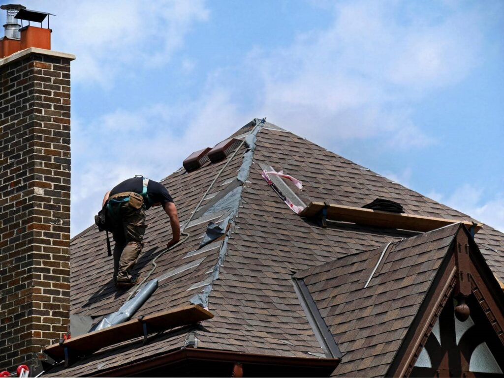 A worker repairs shingles on a steep roof near a brick chimney under a blue sky.