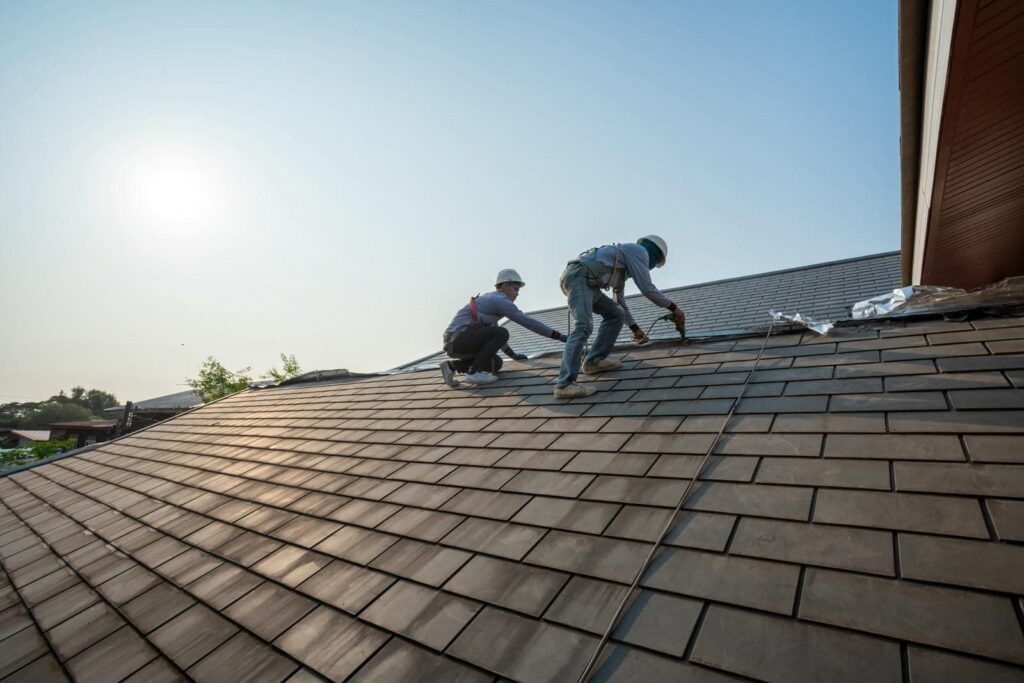 Two construction workers wearing helmets and harnesses install tiles on a sloped roof.