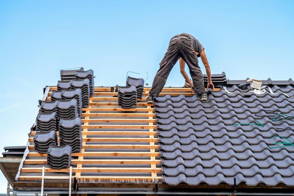 Worker installing dark roof tiles on a wooden roof frame under a clear blue sky.