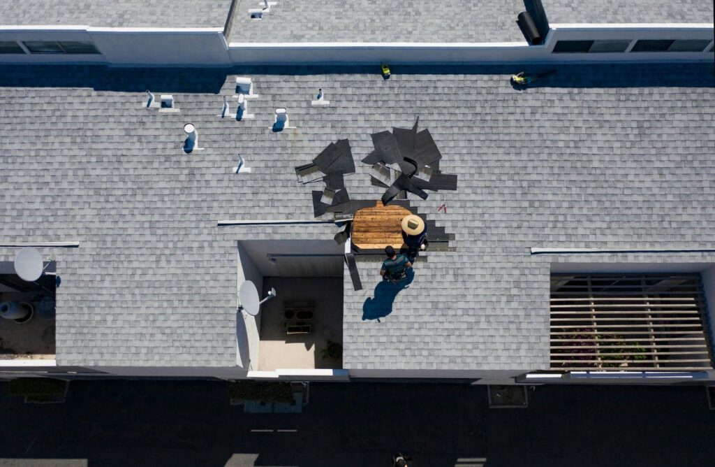 Two workers repairing a roof with exposed wooden planks and scattered shingles.
