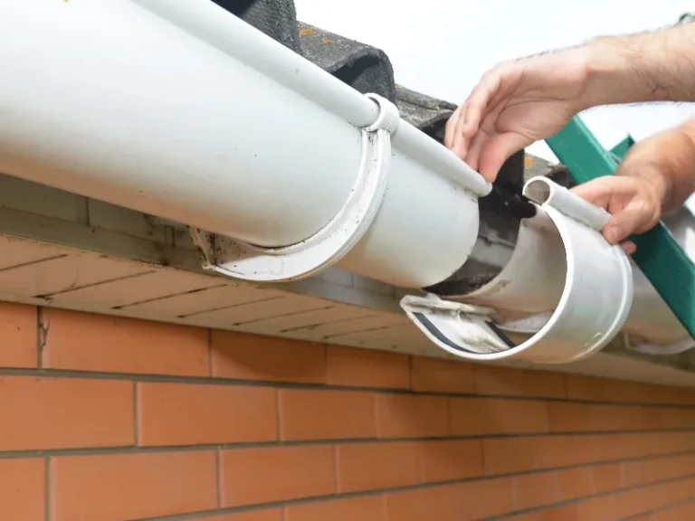 Hands repairing a white rain gutter on a brick house roof edge.