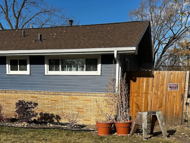 Side of a house with blue siding, brick lower wall, two potted plants, and a wooden gate with a "Beware of Dog" sign.