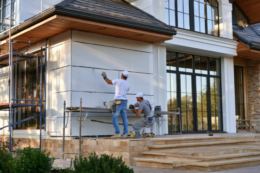 Two workers install siding on a house exterior.