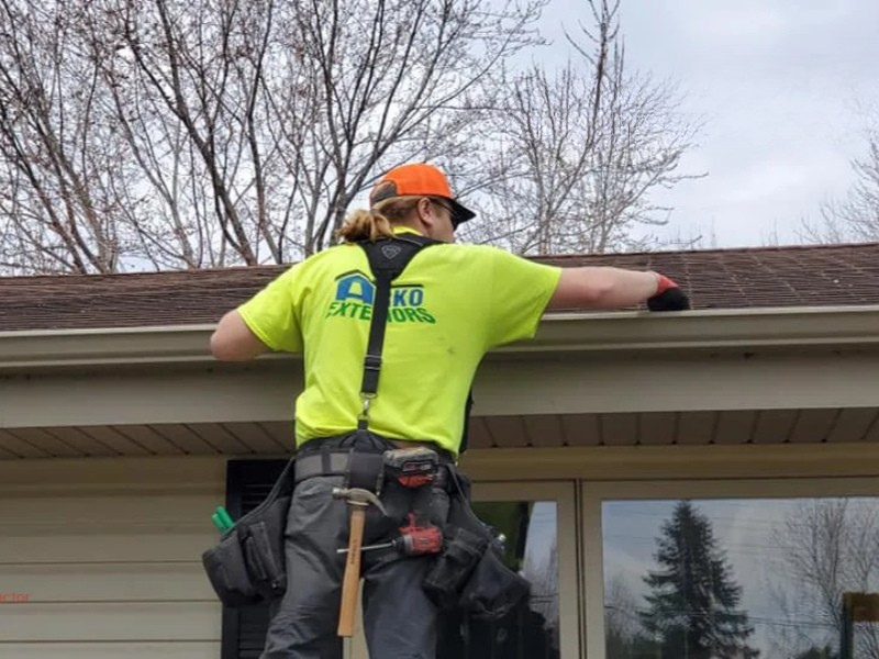 Worker in a neon shirt and orange cap inspecting a roof edge with tools on their belt.