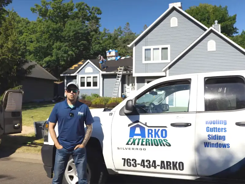 Man in blue shirt and cap stands next to a white ARKO Exteriors truck in front of a house with roofers working.