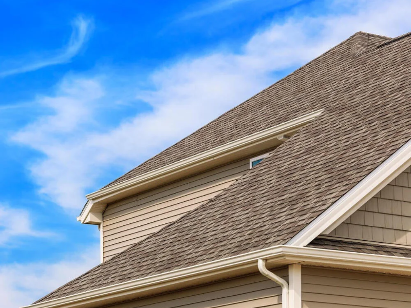 Close-up of a house roof with brown shingles under a partly cloudy blue sky.