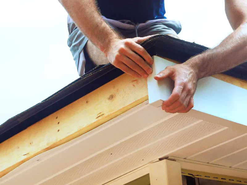 Hands of a person installing white soffit panels under a roof edge.
