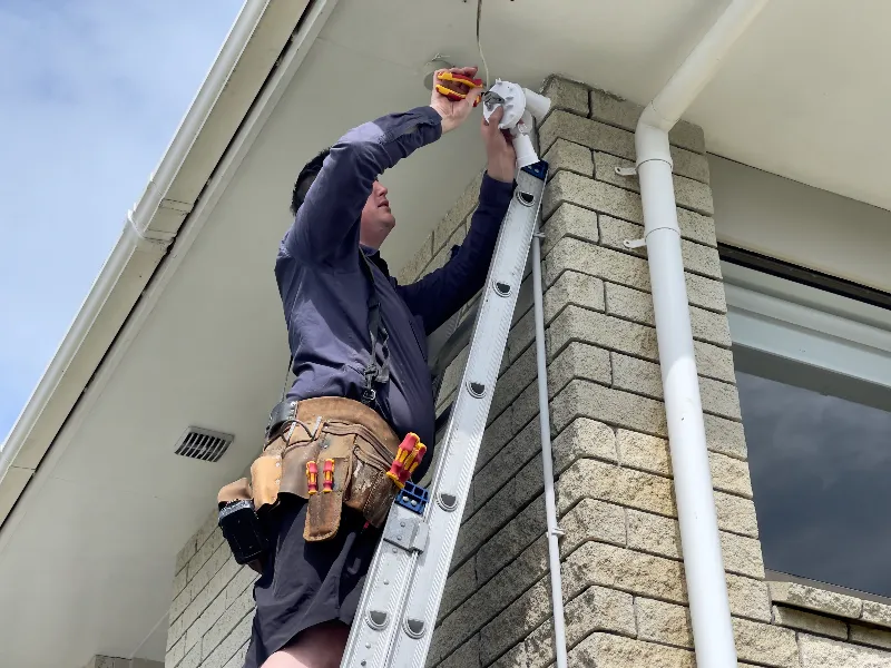 Technician on a ladder installing or repairing a security camera on a building corner.