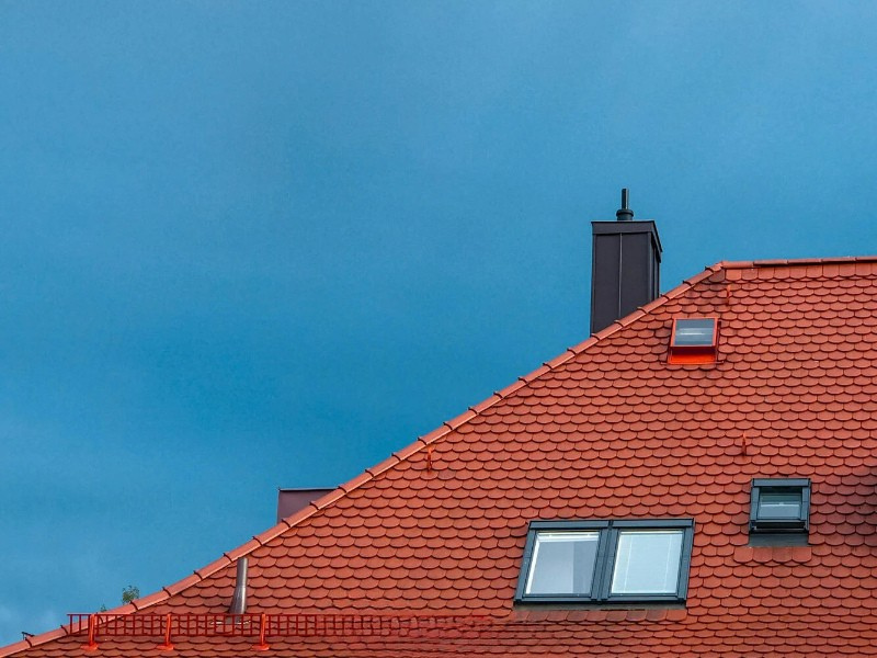 Red tiled roof with three skylight windows and a black chimney against a clear blue sky.
